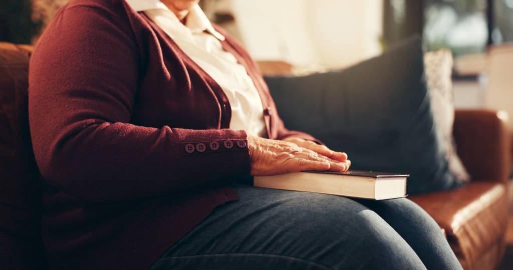 An older woman shoulders down sitting on a couch with Bible on her lap.