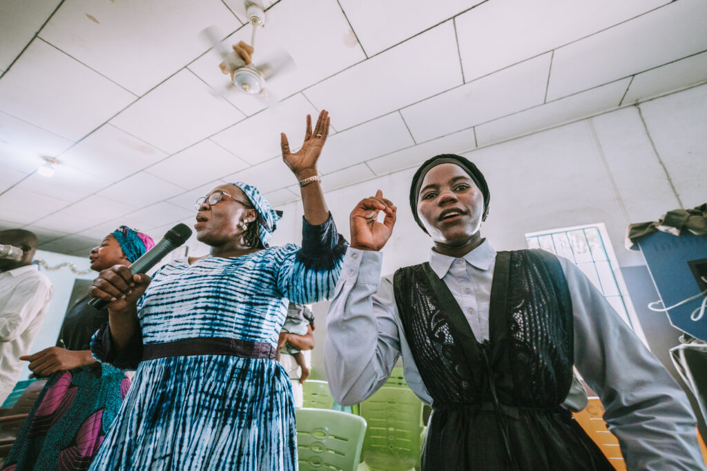 Two West African women lead worship with hands raised in a bright church.