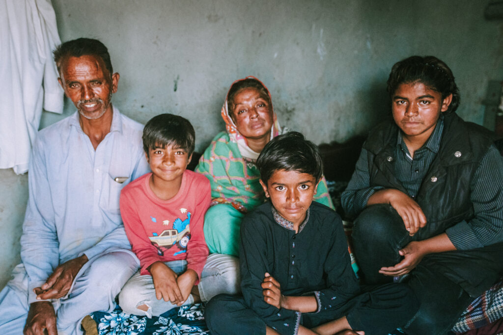 South Asian family seated together in a simple room, looking at the camera with gentle smiles.