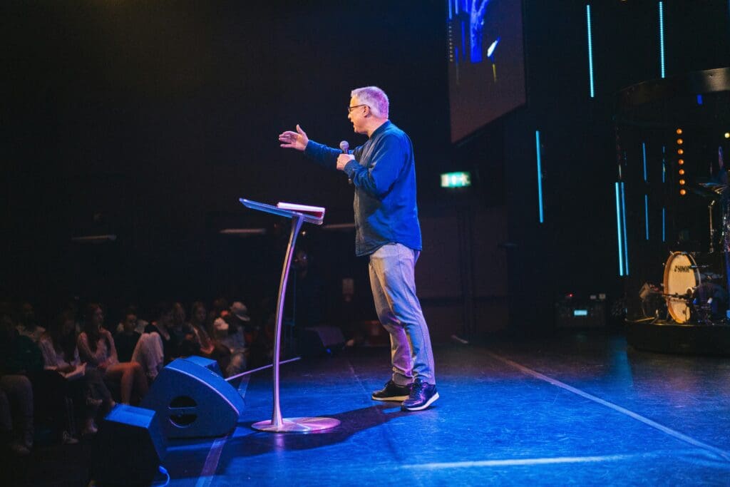 Middle-aged man preaching on stage to a seated congregation in a dimly lit church.
