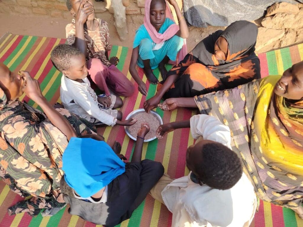 Women and children sit in a circle on colorful striped mats, sharing food from a single bowl.