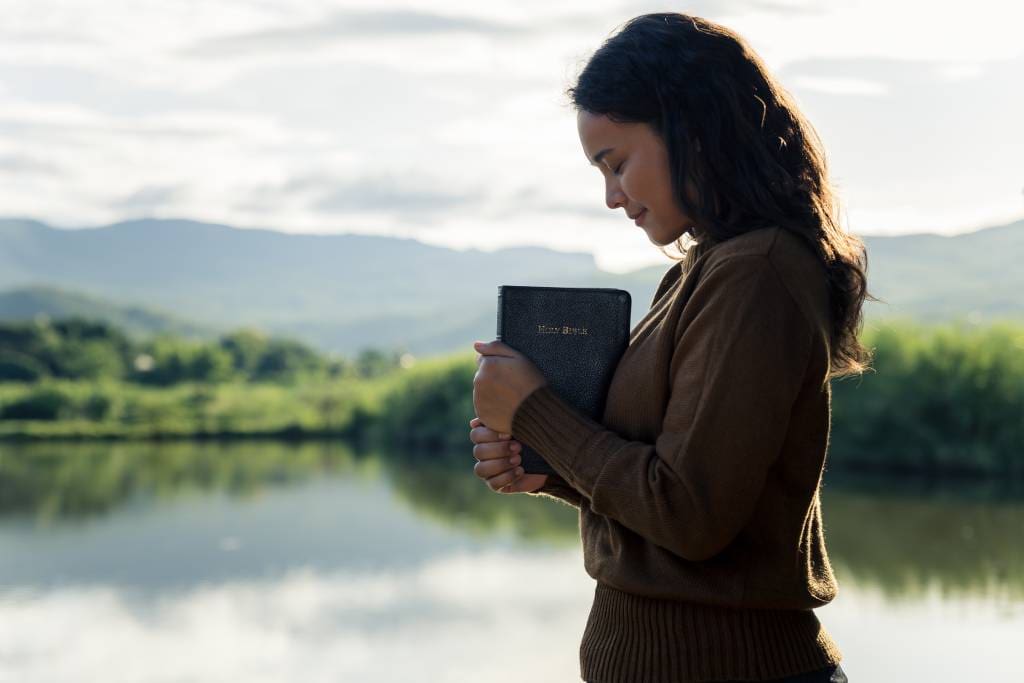 A young woman holding the Bible and praying by a river.