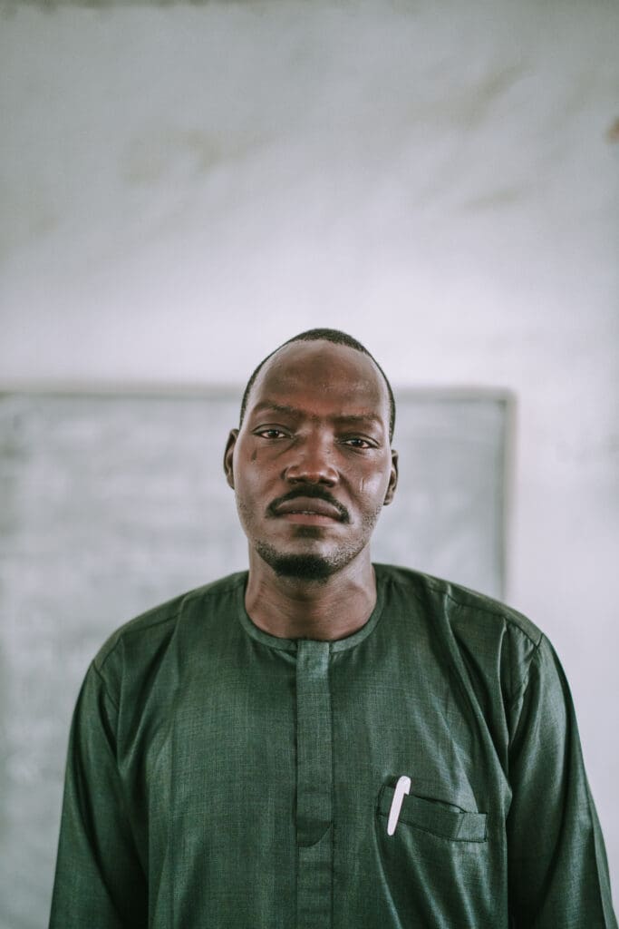A man in a dark green tunic stands before a chalkboard, looking directly at the camera.