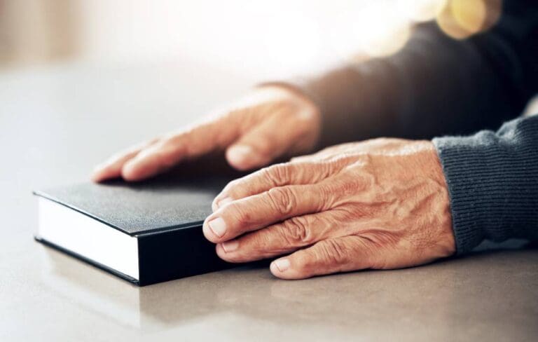 An elderly man’s hands on top of a closed black Bible.