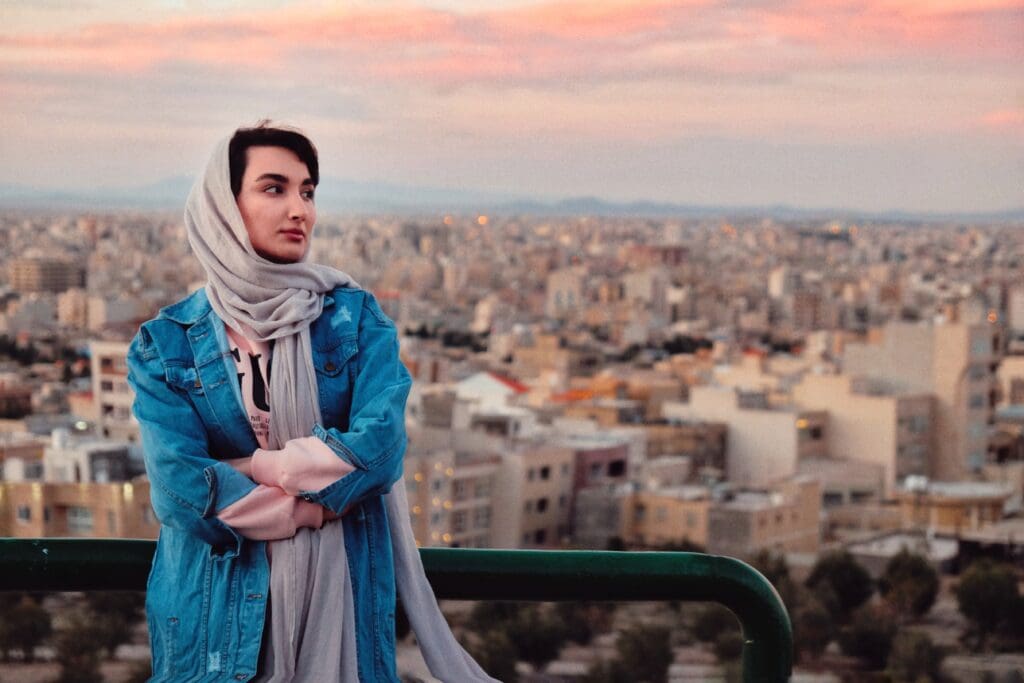 A young woman in a blue denim jacket and gray headscarf stands overlooking a sprawling cityscape at sunset, gazing thoughtfully toward the horizon.