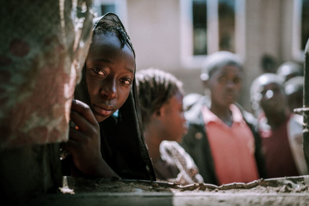 Young Nigerian girl in Plateau State looking toward the camera, with community members visible in the background.