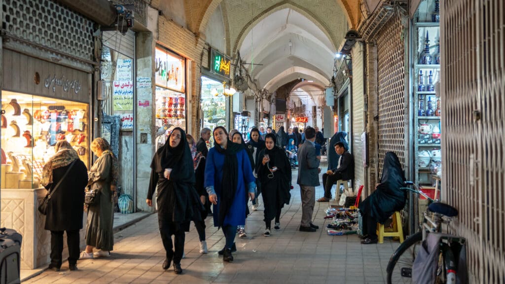 Iranians walking through a traditional covered bazaar with arched ceilings and shop fronts.