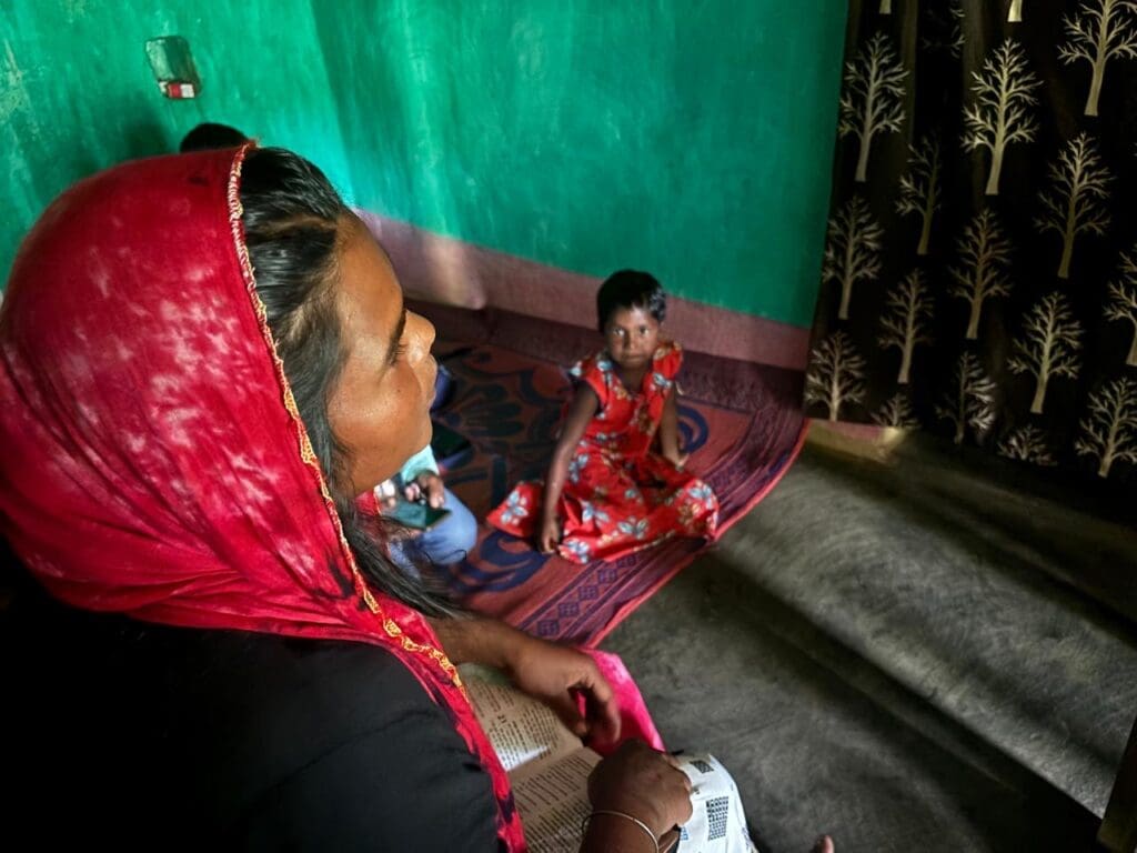 A woman wearing a red head covering sits indoors with an open Bible in her lap, while a young girl sits behind her.